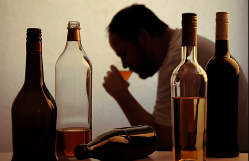5 sticle cu alcool asezate pe o masa, cu o persoana de gen masculin in plan secund care bea dintr-un pahar