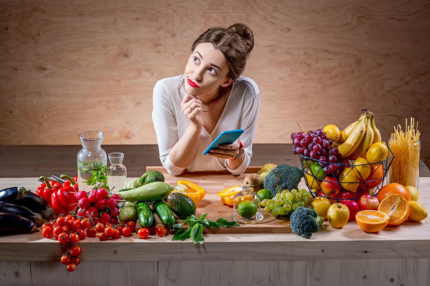 femeie care se gandeste cu telefonul in mana si este inconjurata de multe fructe si legume
