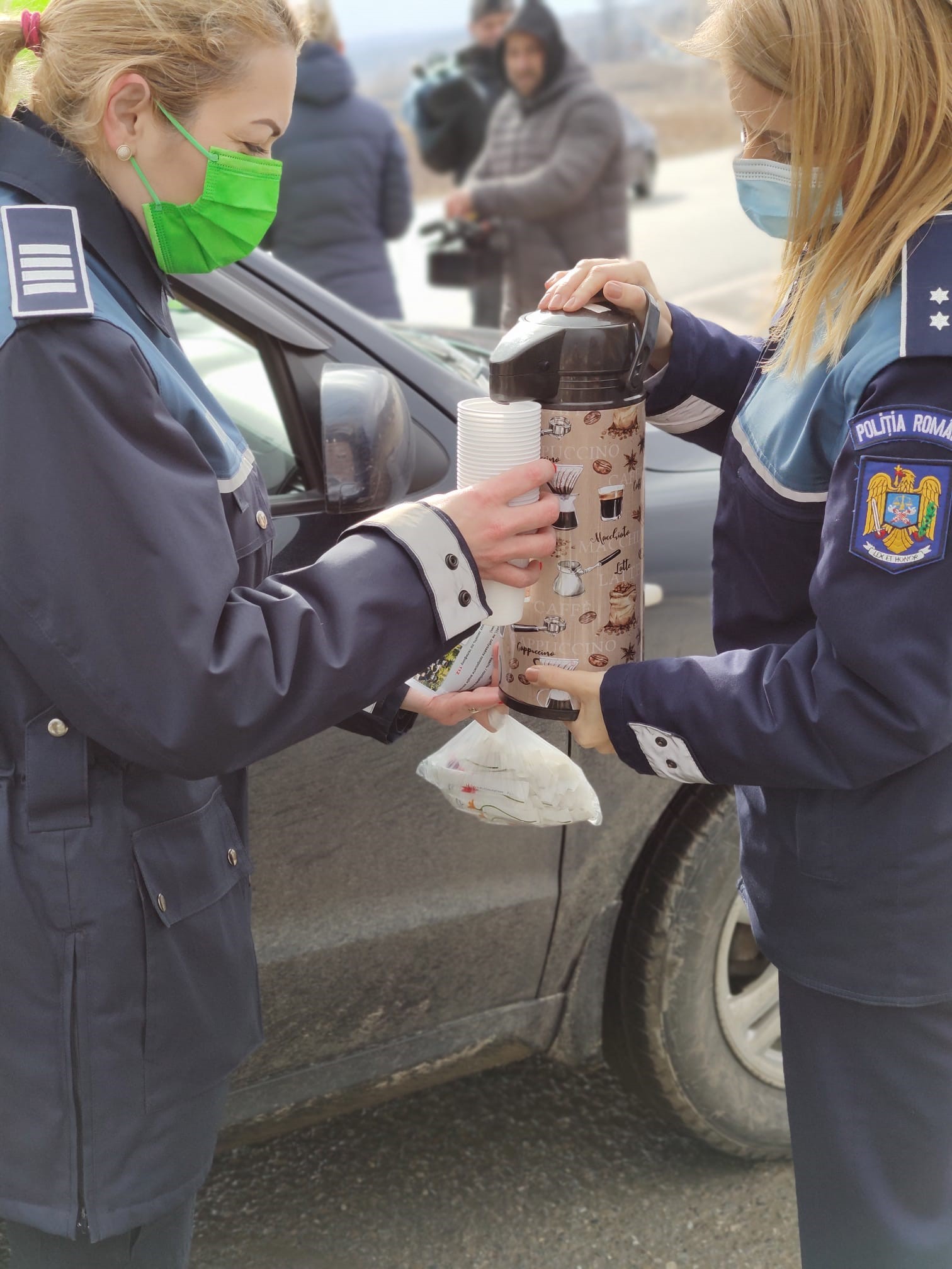 Doua femei imbracate in uniforma Politiei Romane toarna ceai dintr-un termos langa o masina neagra