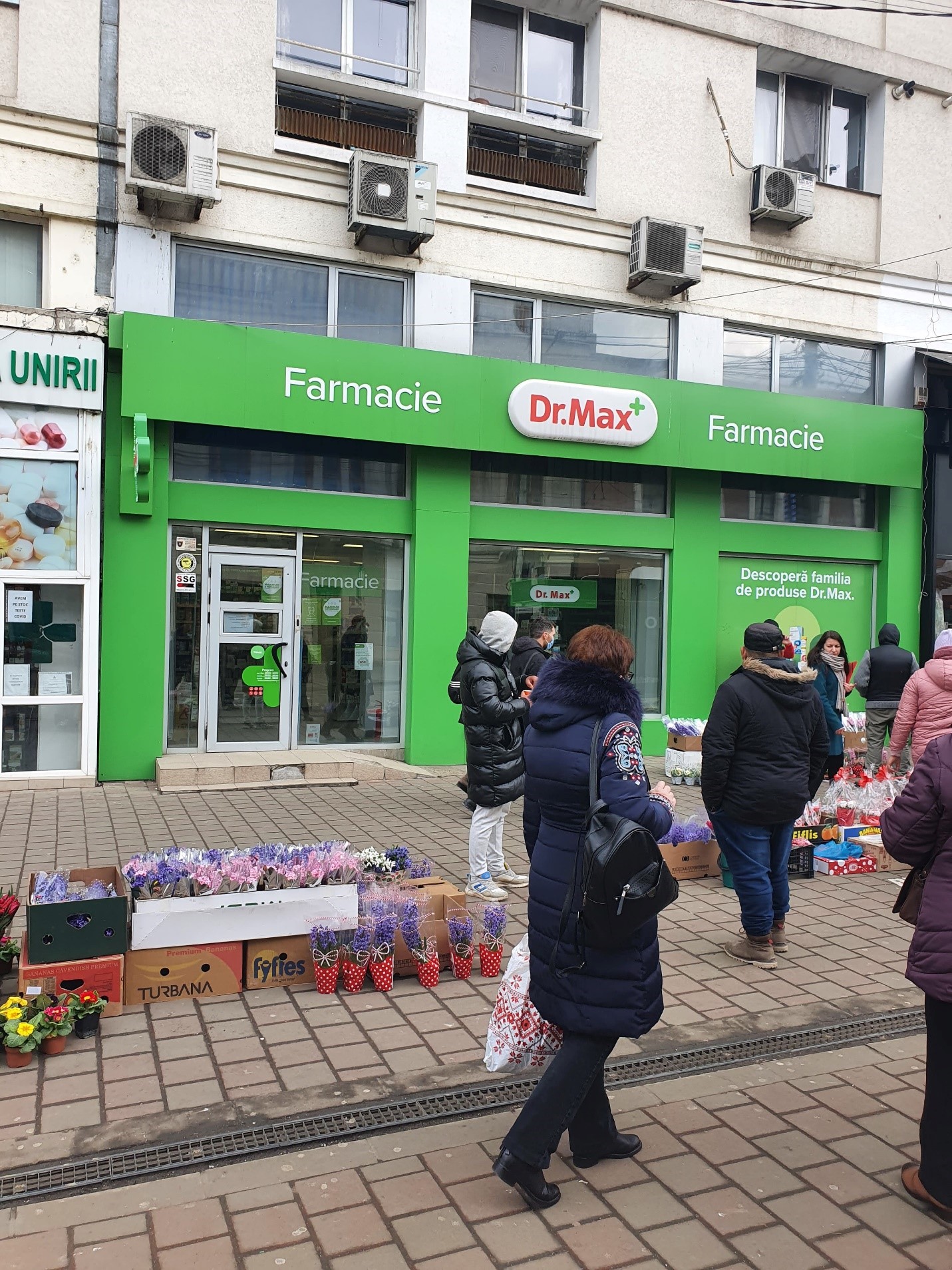 O farmacie Dr. Max in fata caruia se vad cateva cutii cu flori si cateva persoane care asteapta tramvaiul