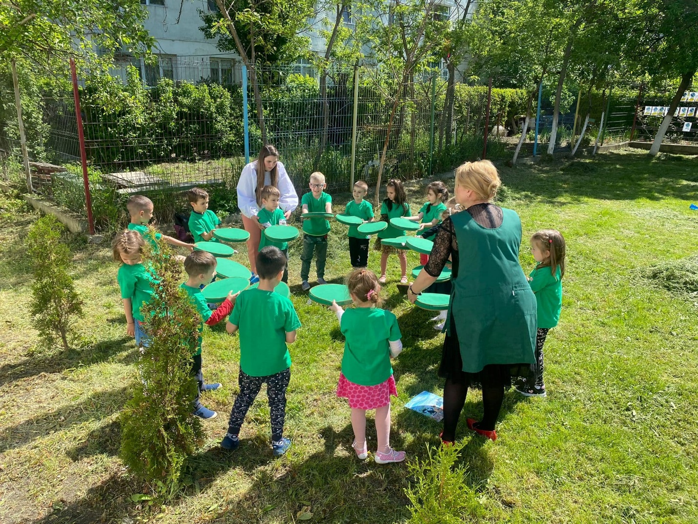 copii asezati in cerc, imbracati in verde, si doua persoane adulte care ii coordoneaza