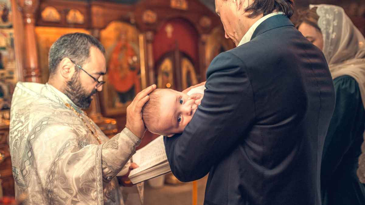 nasi care tin in brate un bebelus in fata unui preot care citeste din biblie